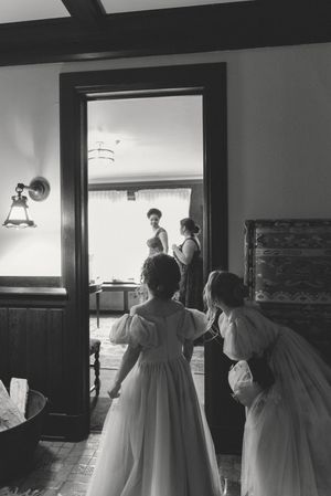 Black and white photo of flower girls in white dresses peering through a doorway at bridesmaids during wedding preparations.