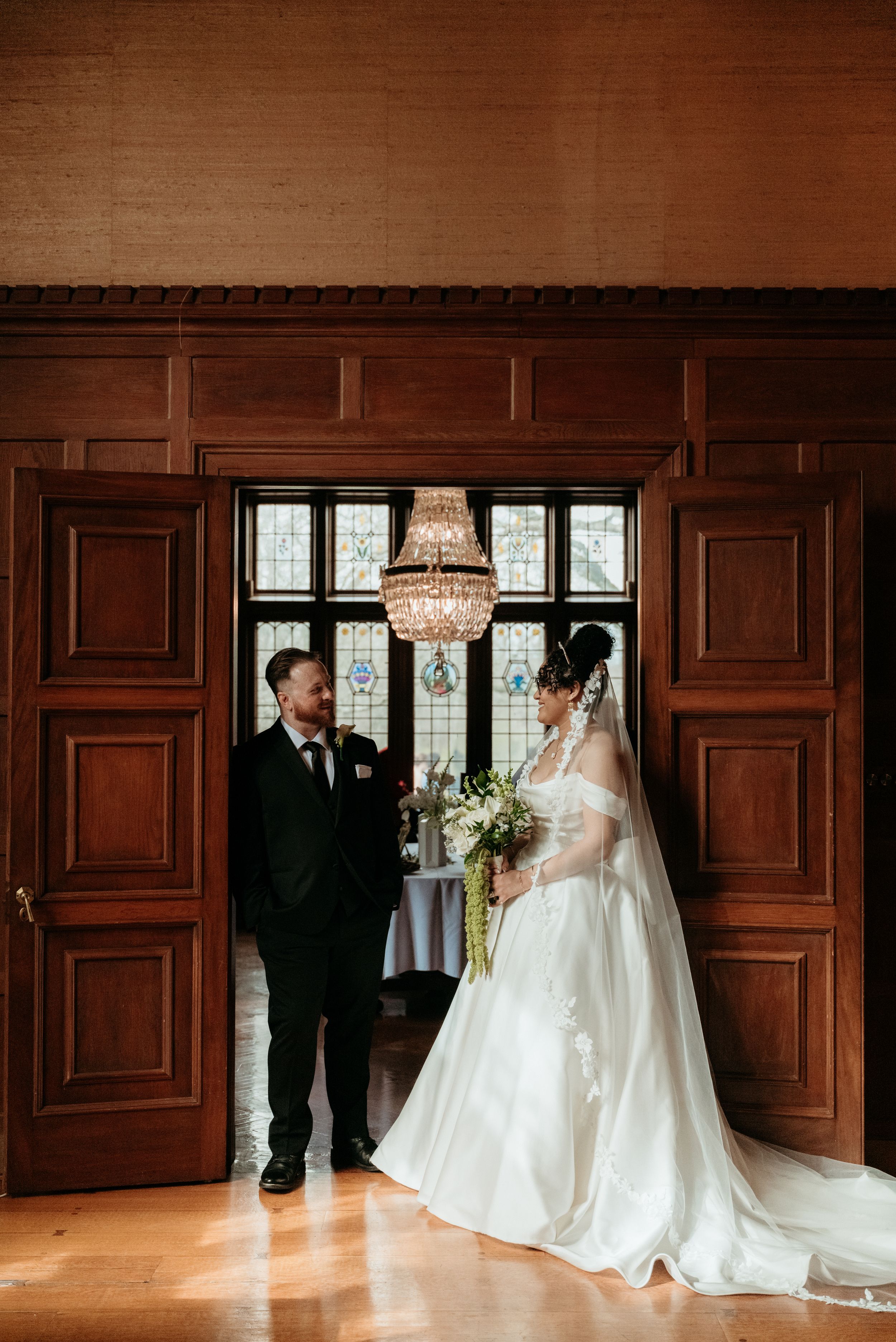 Bride and groom share a first look moment in an elegant wood-paneled room with a crystal chandelier.