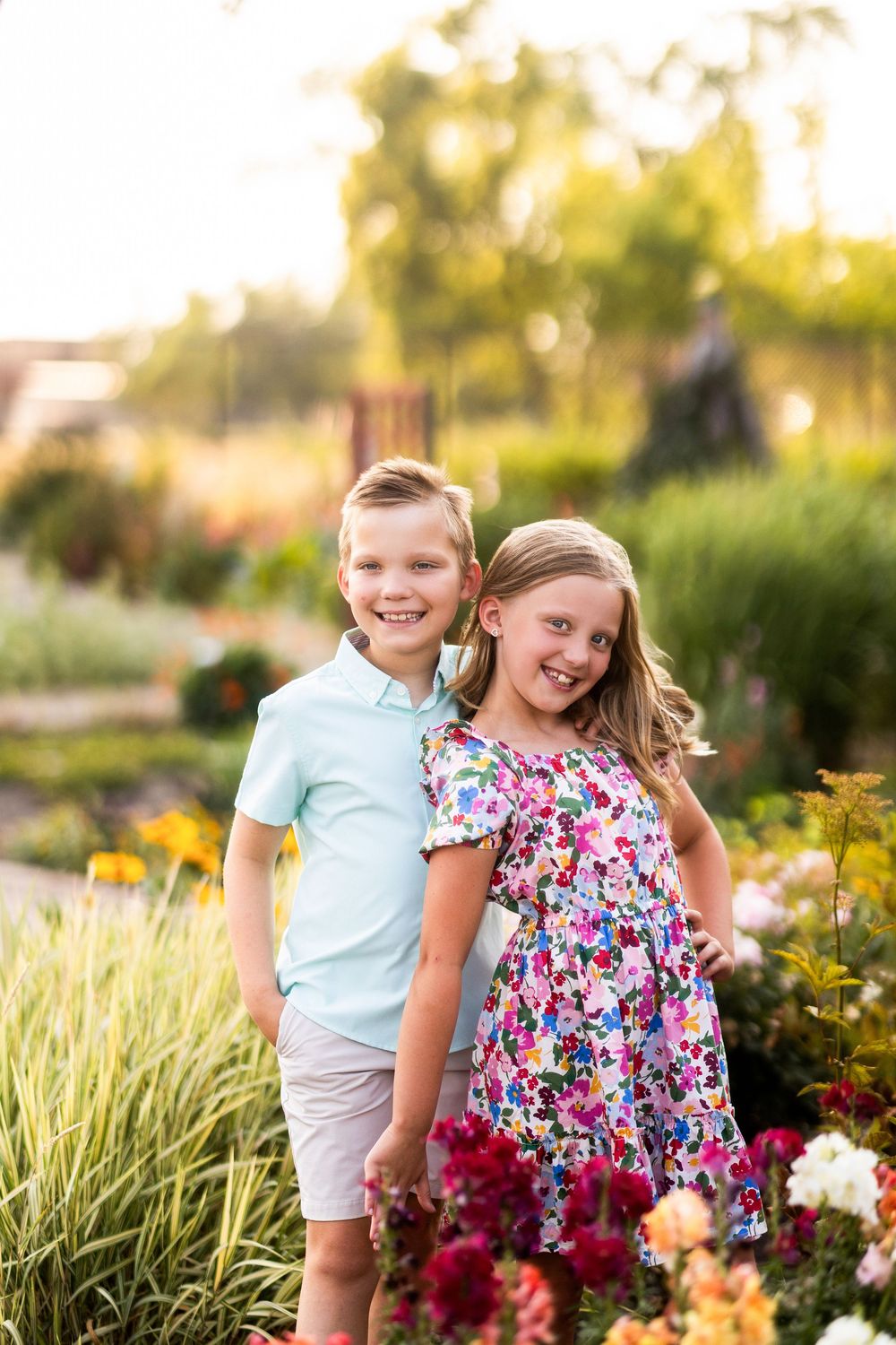 Two smiling children pose together in a colorful garden, girl in floral dress and boy in blue polo shirt.