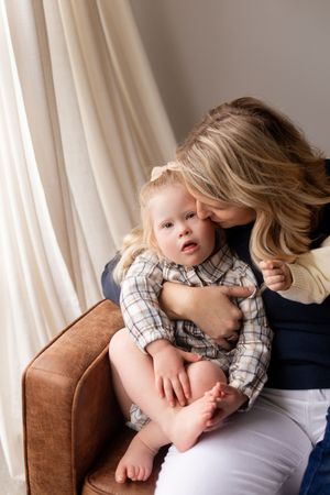 A tender moment captured between parent and baby sitting on a brown leather chair by a curtained window.