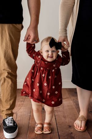 A baby in a red floral dress stands between adult feet on a hardwood floor.