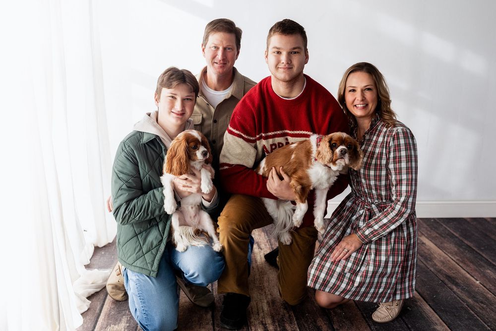 Family group sitting on wooden floor with three adorable Cavalier King Charles Spaniel puppies.