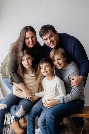 A family of five sits close together smiling warmly in a casual indoor portrait against a light background.