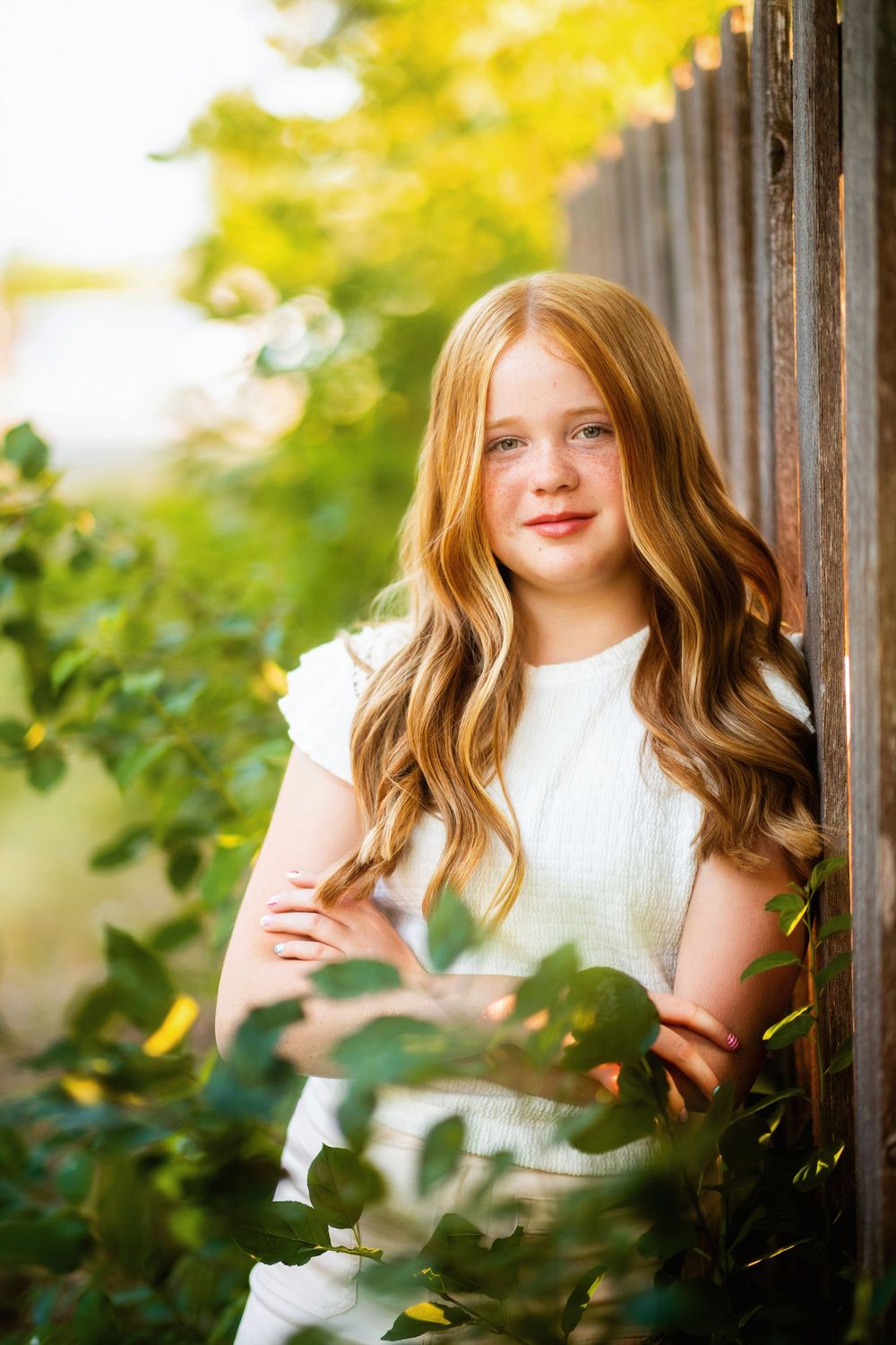 Young girl with long wavy red hair in white dress leaning against wooden fence surrounded by green foliage.