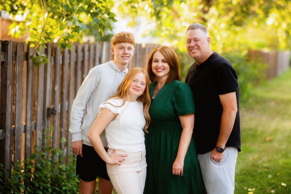 Family of four smiling together outdoors in a lush green backyard, wearing casual and elegant attire.
