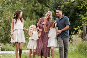A family walks together through a lush garden setting wearing coordinated white and gray outfits.