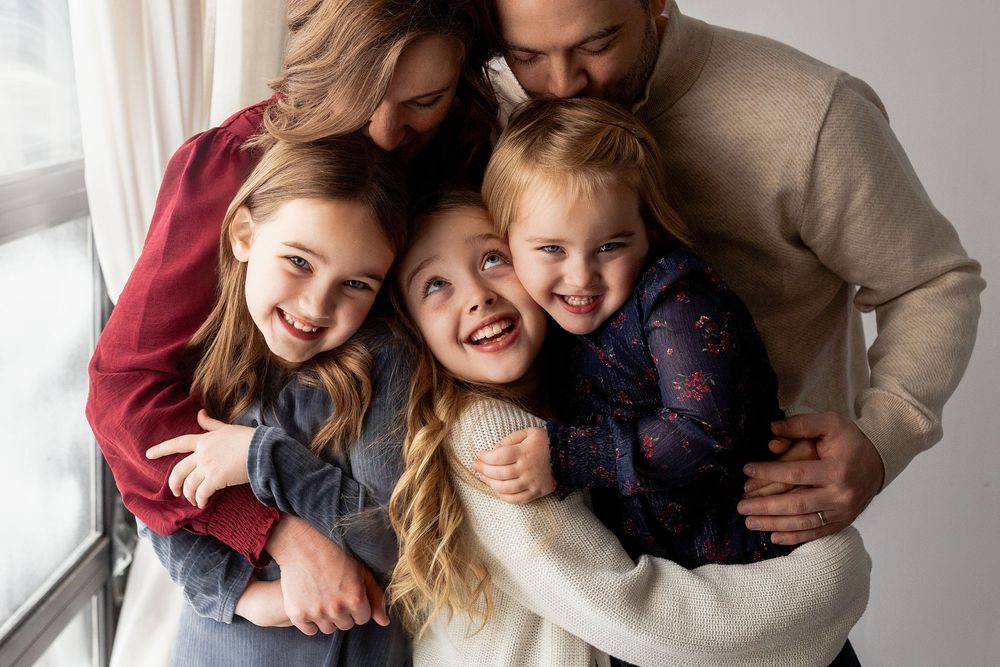A joyful family embraces and laughs together during an indoor photo session with matching casual outfits.