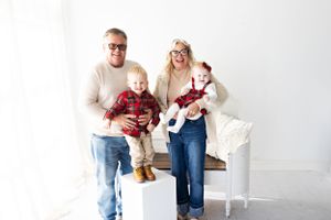 Family of four in coordinating outfits posing on white furniture against bright background.