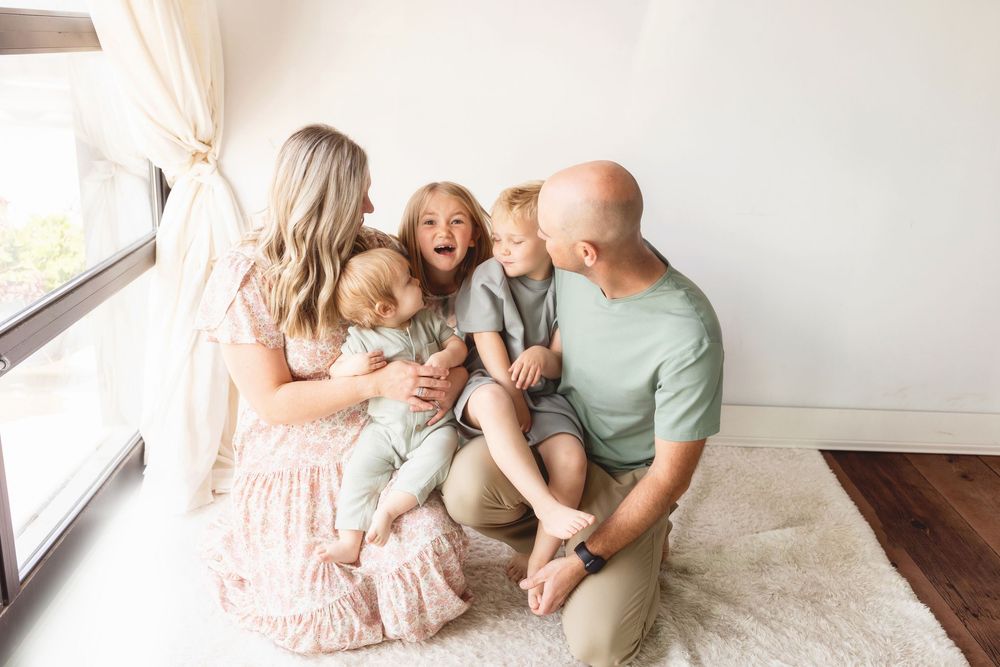 A family shares joyful laughter while sitting together on a light carpet near a window with white curtains.