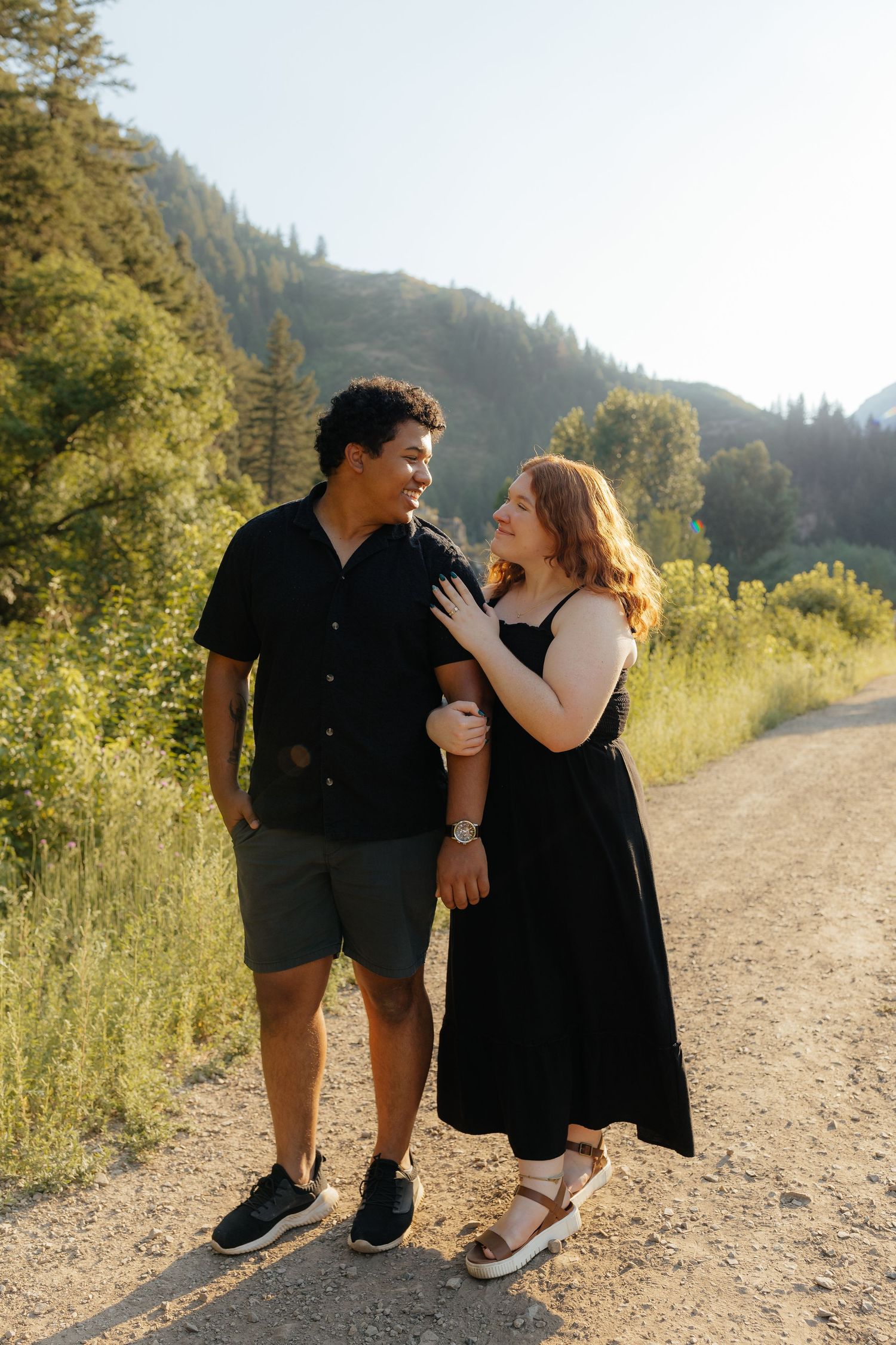 Interracial couple smiling at each other on a sunny mountain dirt path.