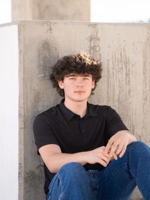 A young person in a black polo shirt and jeans sits casually against a concrete wall in an outdoor portrait setting, perfect for a senior