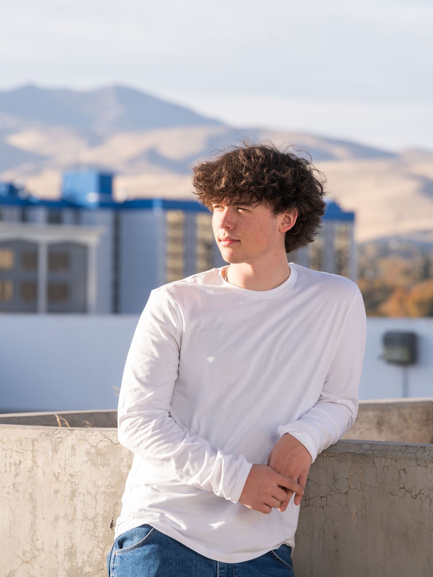 Person in white long sleeve shirt and jeans poses against concrete wall with mountains and buildings in background.
