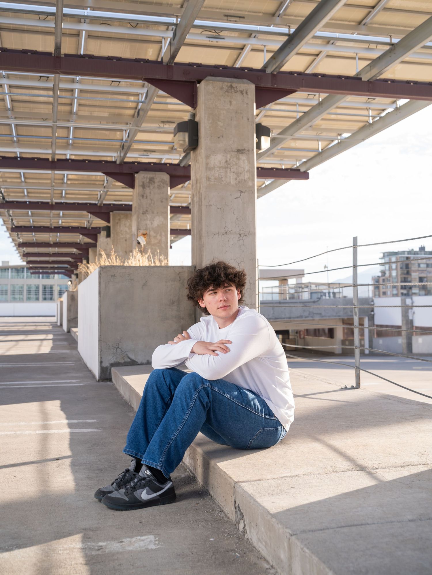 A person in a white shirt and jeans sits casually on concrete steps under an industrial covered walkway at sunset.