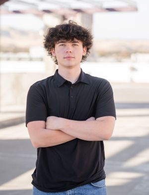 High school senior in a black polo shirt and jeans stands with crossed arms against a blurred outdoor background.