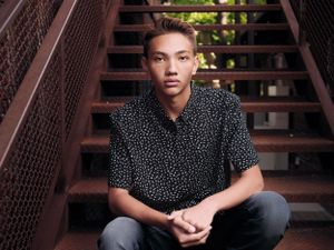 Youth wearing a black bold patterned shirt sits on rustic metal stairs with hands clasped in casual portrait pose.