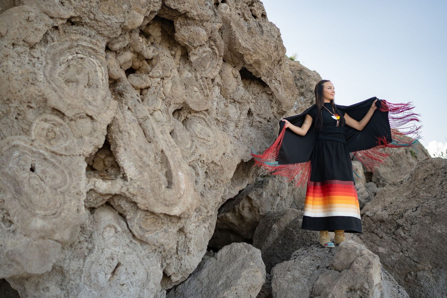 A black dress with rainbow gradient trim flows dramatically against a rocky cliff face at sunset.