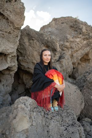 A person wrapped in a black and red fringed shawl sits among rugged rock formations in an outdoor setting.