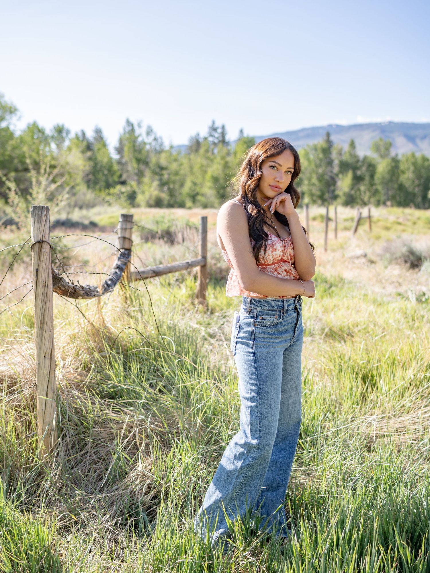 Casual country style portrait in a rural field with wooden fence posts and mountains in the background on a sunny day.