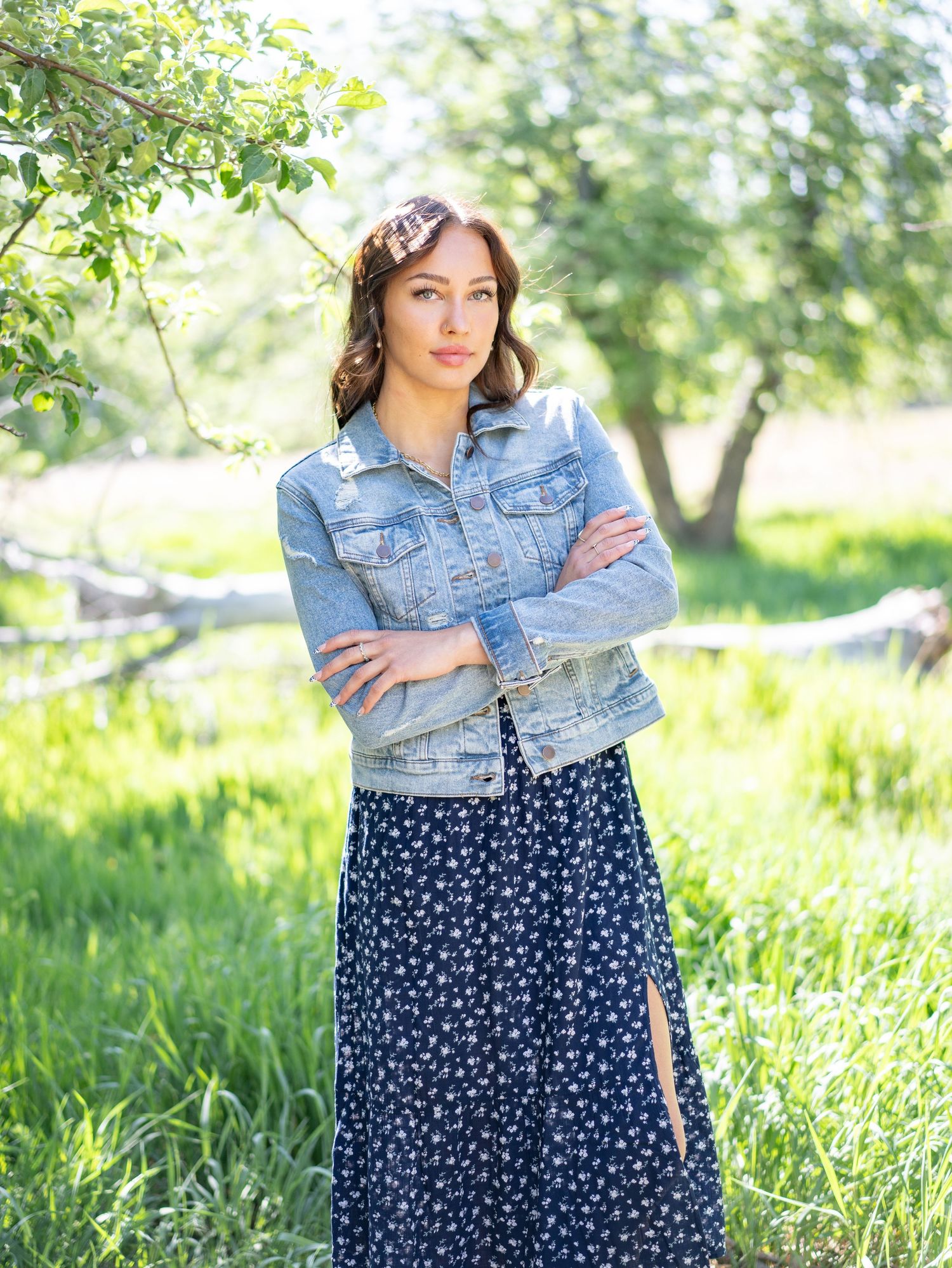 Senior girl wearing a denim jacket and floral maxi skirt poses outdoors in a sunlit garden setting.