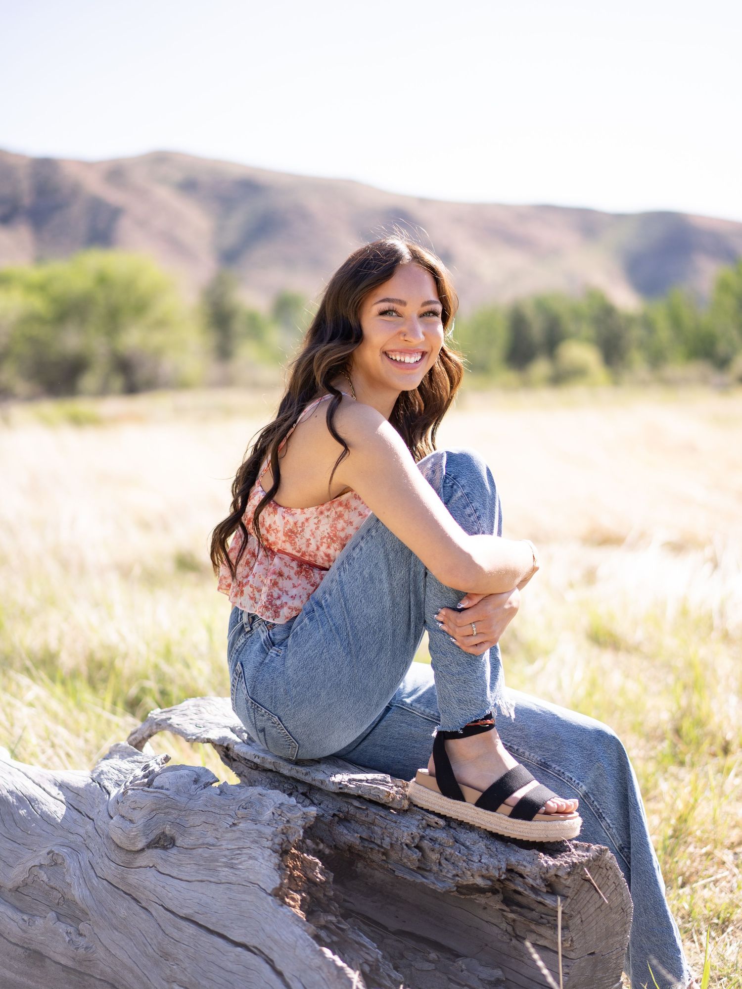 A high school senior wearing jeans, floral shirt and sandals sits on a large stump in a sunny field with mountains in the background.