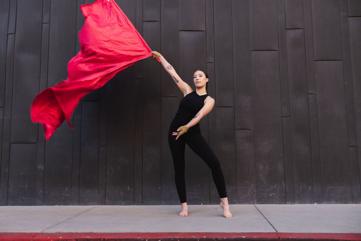 High school senior in black outfit performs dynamic movement with flowing red fabric against dark wall background.