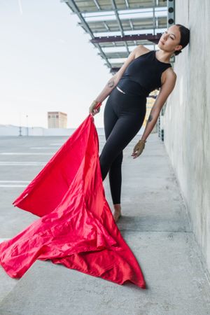 Athletic dancer in black workout attire poses gracefully with flowing red color guard flag in a modern urban setting.