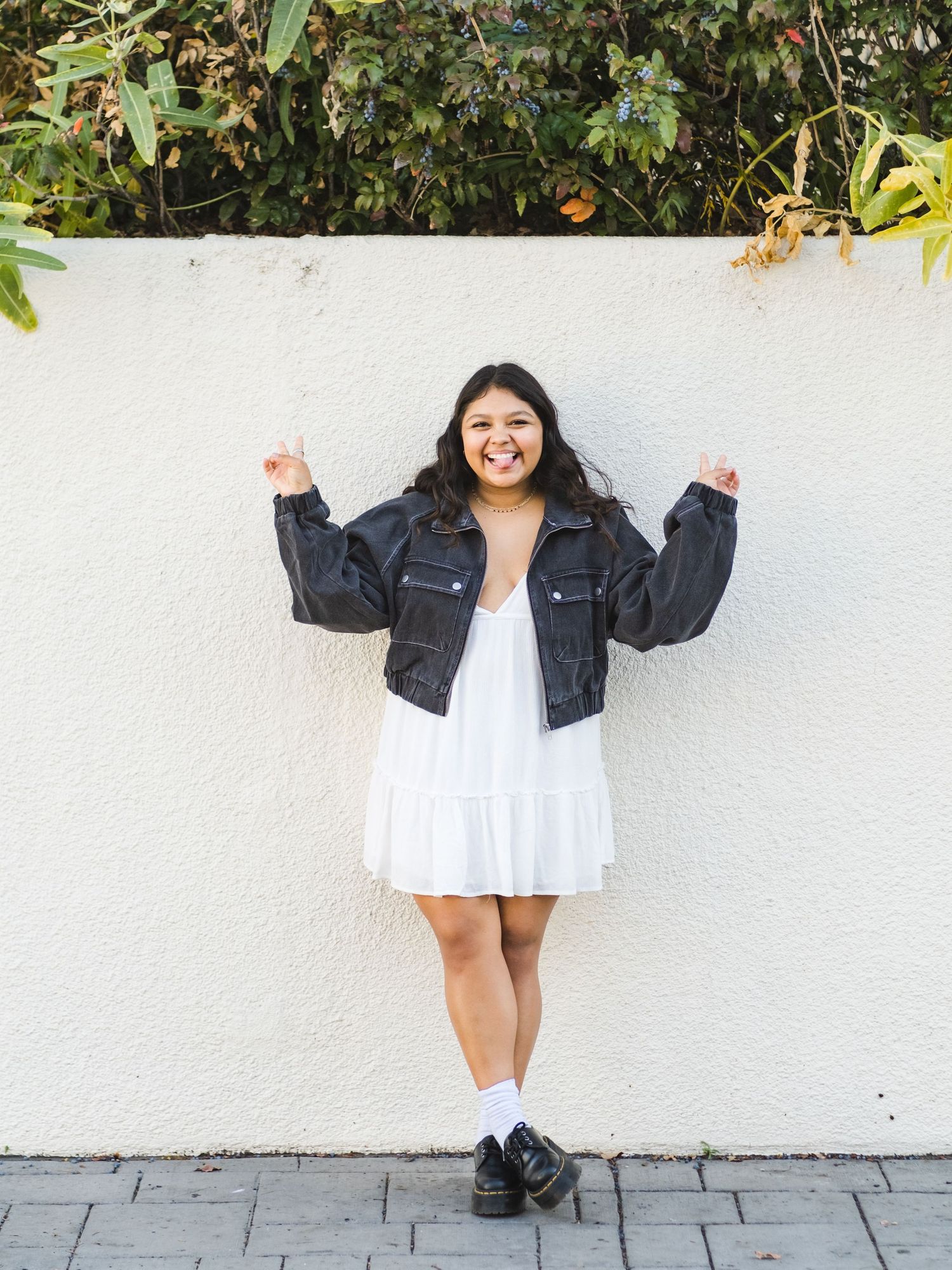 High school girl in a black denim jacket and white dress poses playfully against a white wall with plants in the background.