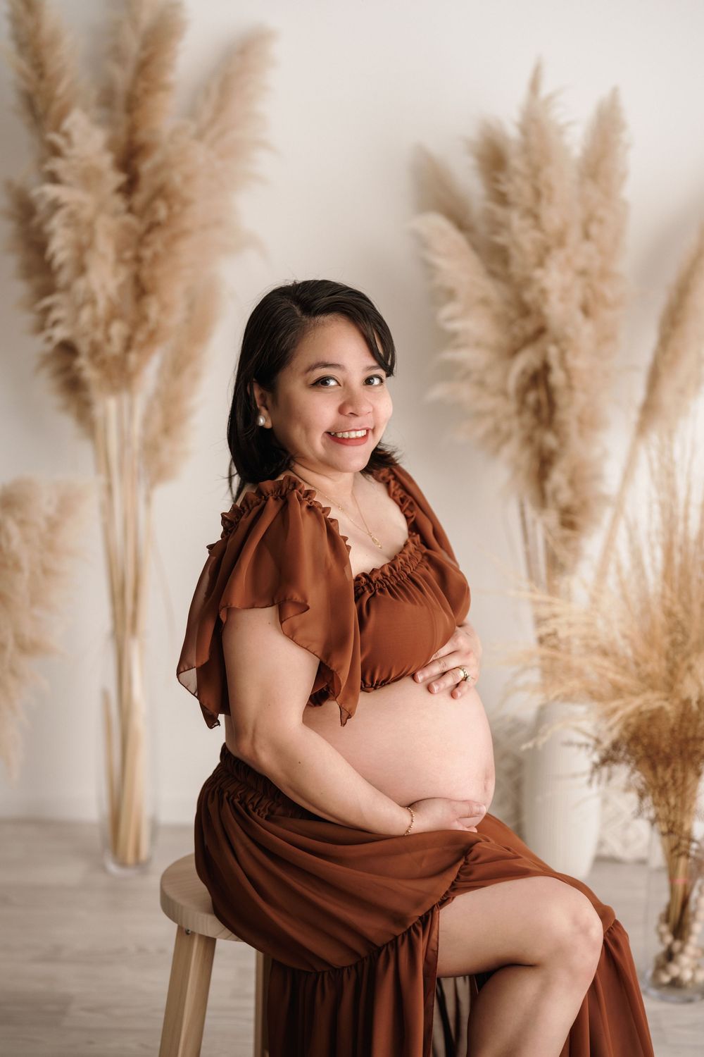 Expectant mother in brown dress seated elegantly against backdrop of pampas grass in light studio setting.