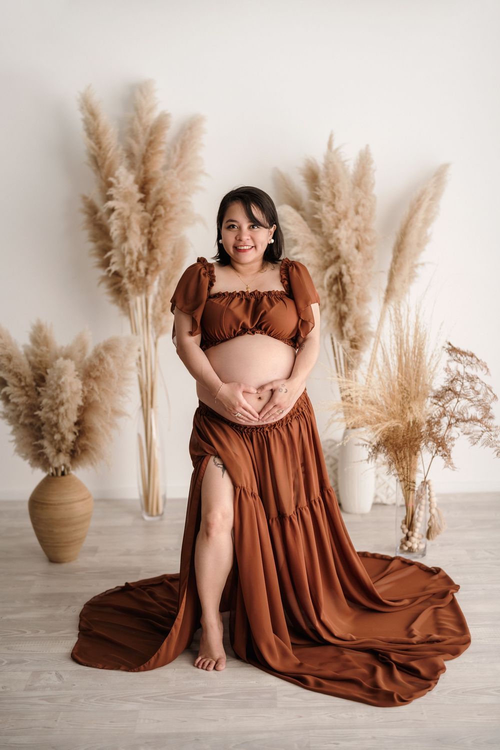Maternity photo featuring a flowing brown dress and decorative pampas grass arrangements in a minimalist studio setting.
