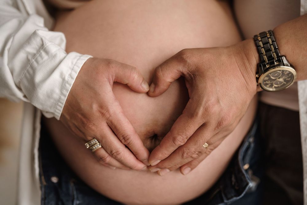 Hands form a heart shape over a pregnant belly while wearing a silver watch and engagement ring.