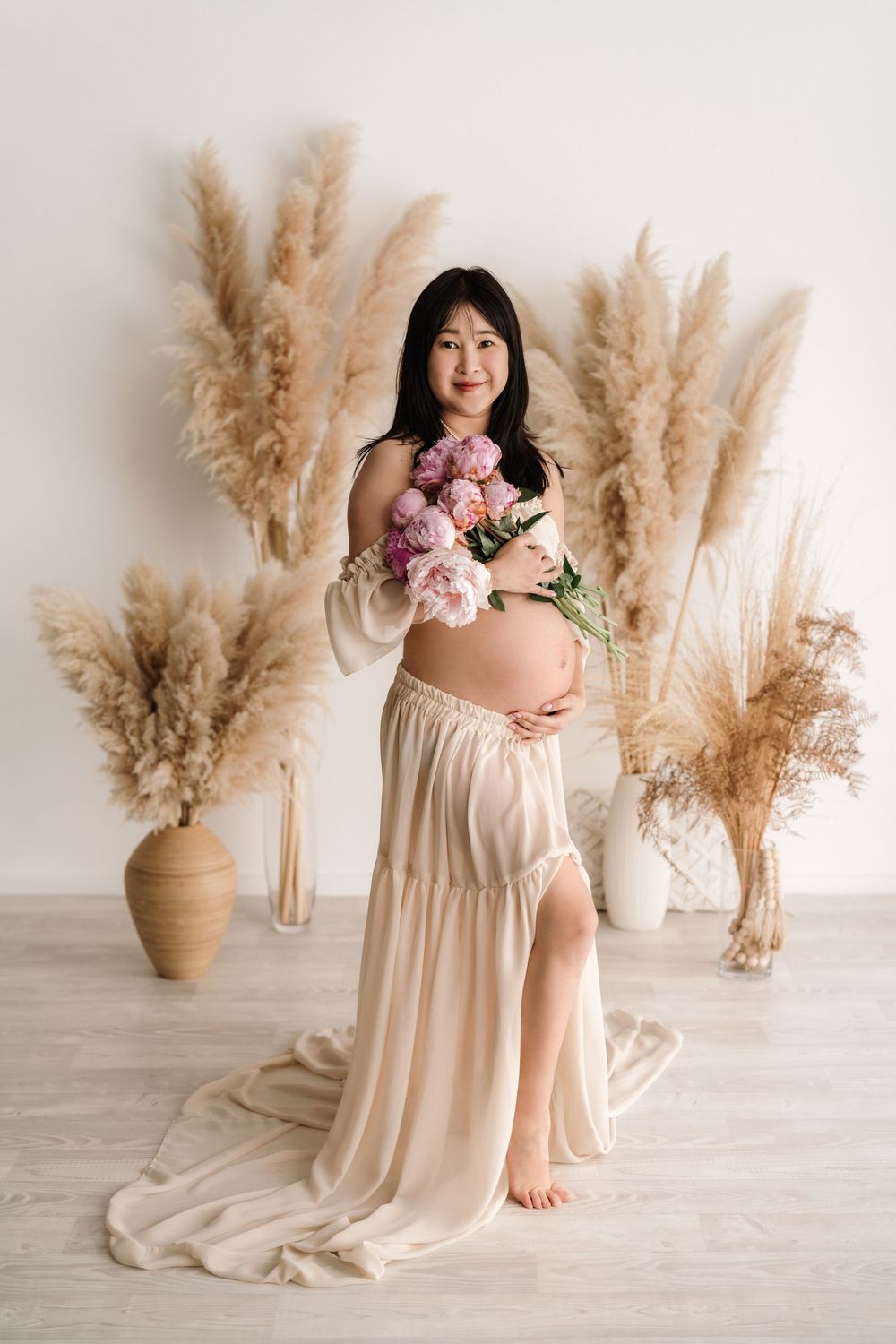 Maternity photograph featuring pink flowers and pampas grass decor in a light, airy studio setting.