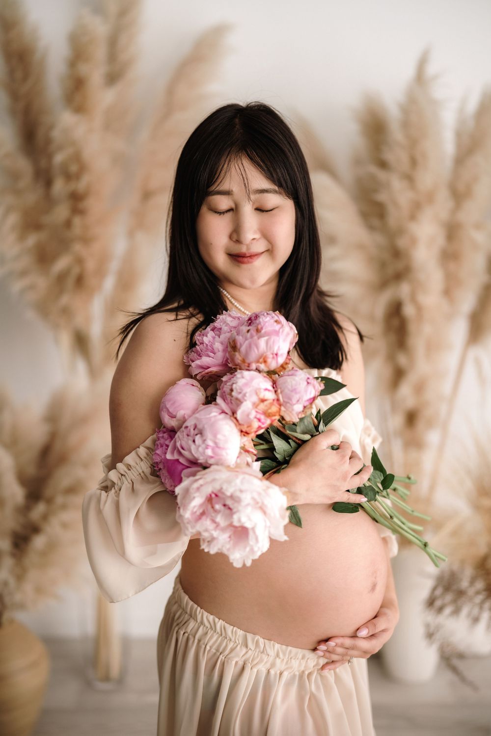 Expecting mother in flowing dress holds pink peonies while posing against pampas grass backdrop during maternity photoshoot.