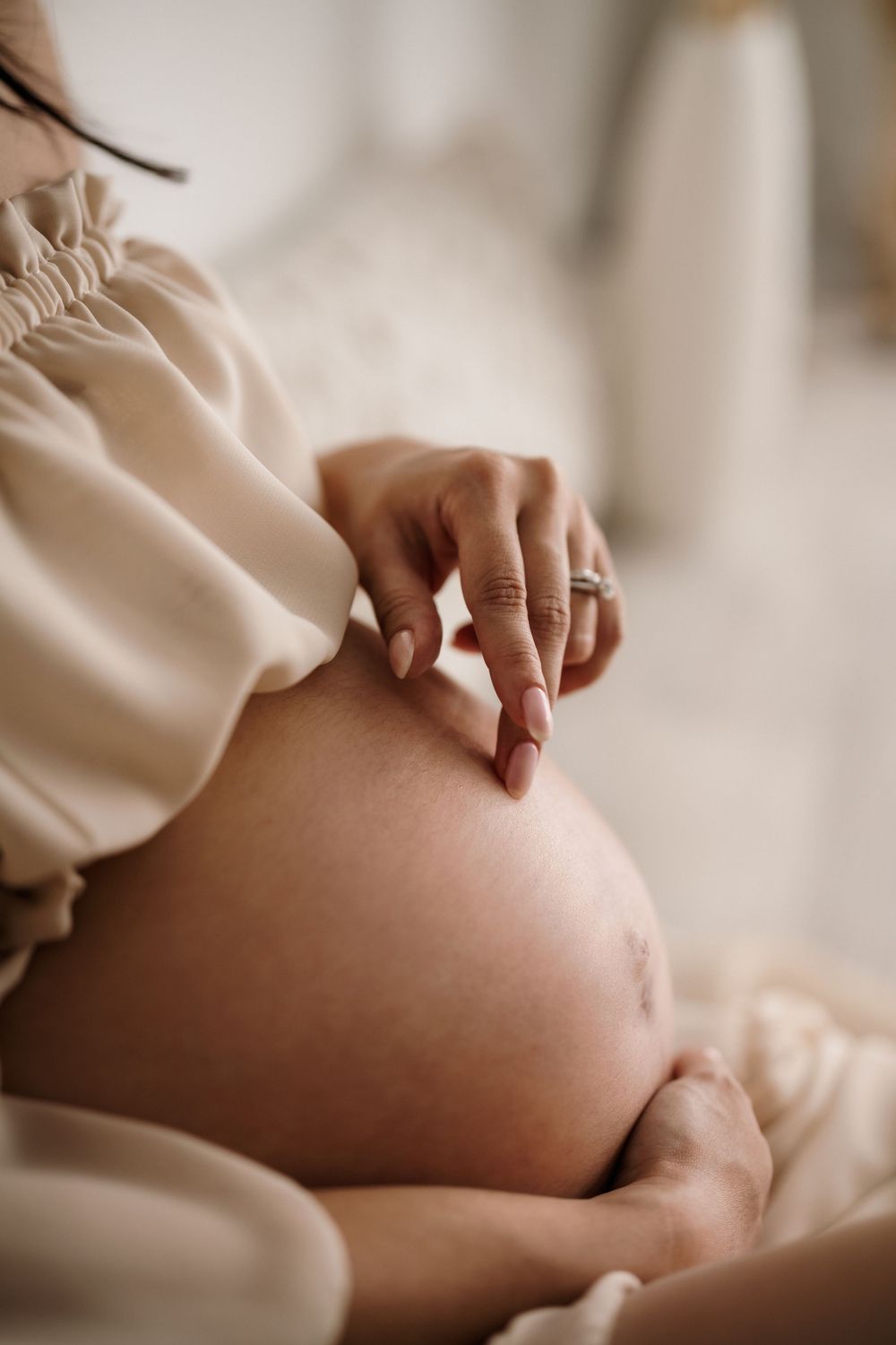 A gentle hand rests on a pregnant belly while wearing a cream-colored flowing dress in soft lighting.