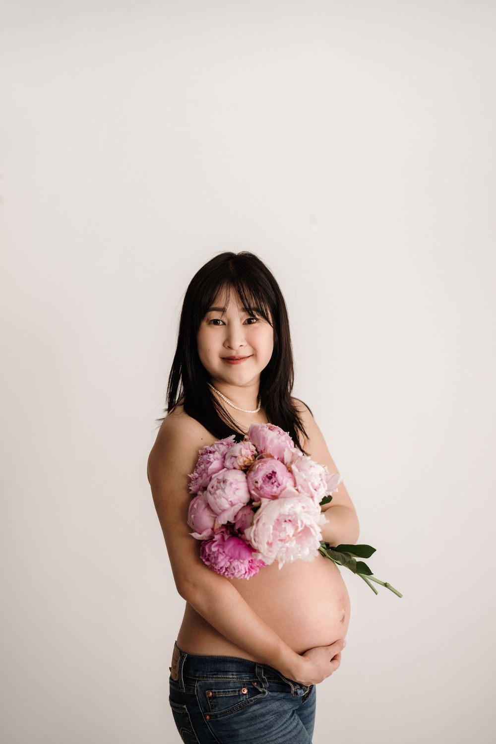 Side view of an expecting mother in jeans holding a bouquet of pink and purple peonies against a light background.