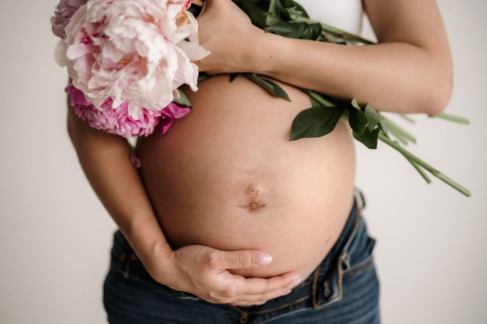 A pregnant belly is gently cradled while holding a pink peony flower against a neutral background.