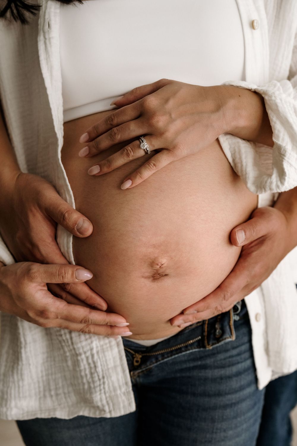 Close up of pregnant belly with hands wearing engagement ring touching bare stomach in white shirt and jeans.