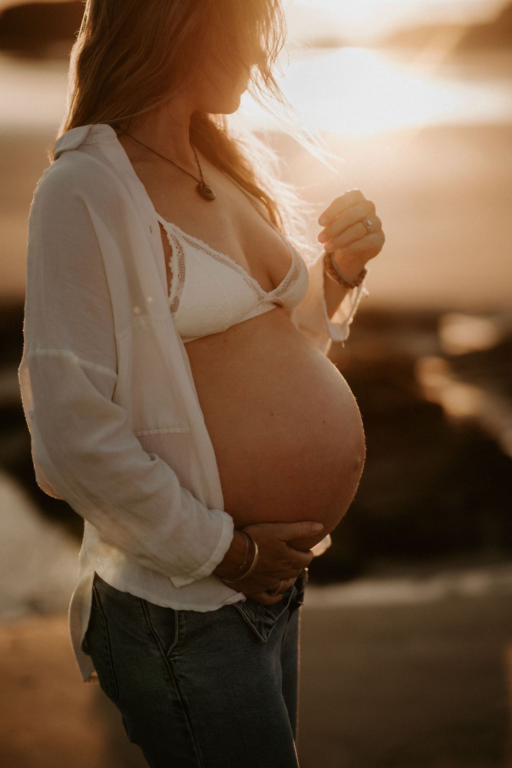 Silhouetted pregnant woman in white shirt and jeans stands in warm sunset light holding her belly.