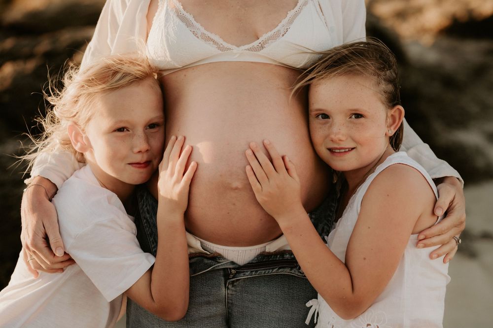 Two children in white shirts embrace and touch a pregnant woman's bare belly in a warm sunlit outdoor setting.