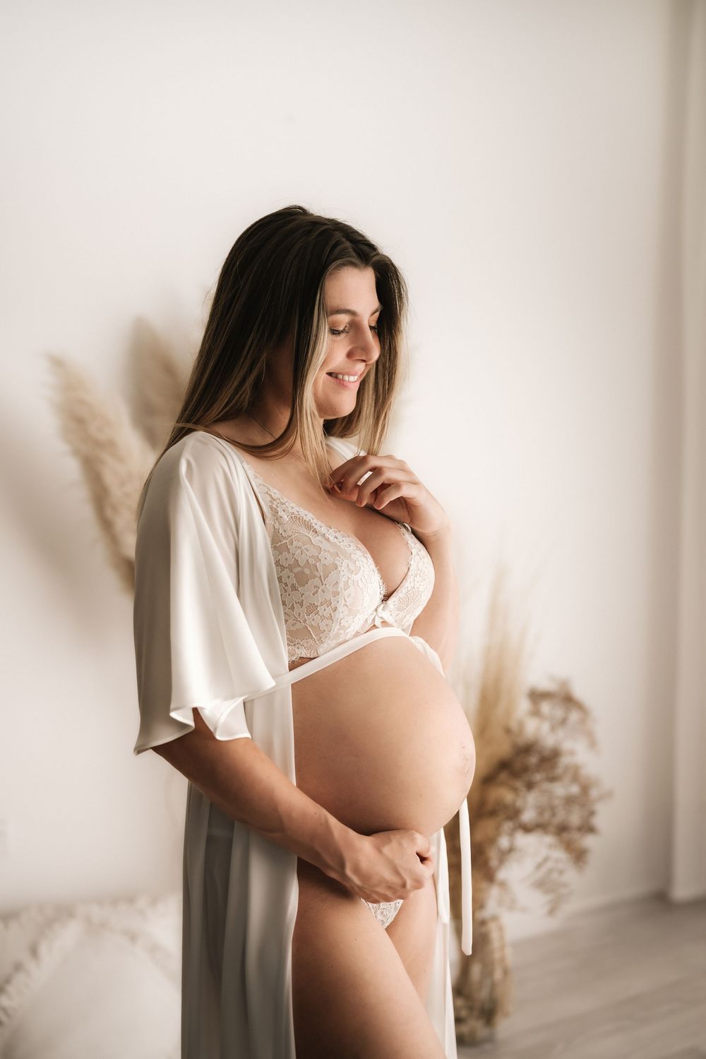 Expectant mother in white robe and lingerie poses against neutral wall with dried pampas grass decor.