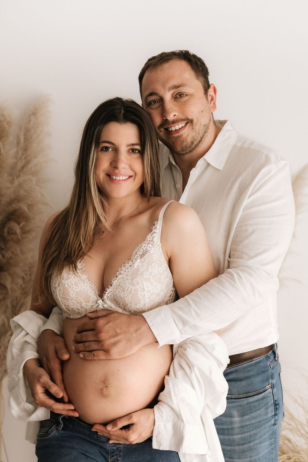A loving couple poses together for a maternity photo against a neutral backdrop with pampas grass decor.