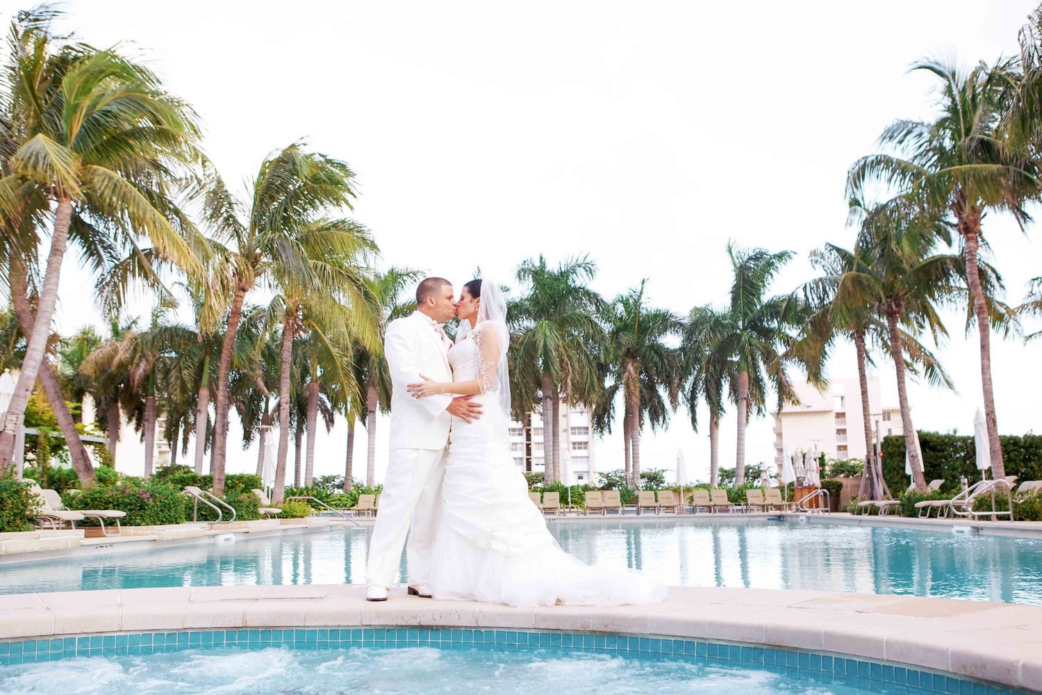 A couple in white attire stands by a tropical resort pool surrounded by tall palm trees on a sunny day.