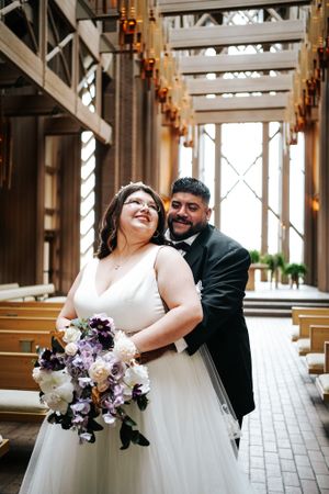 A joyful wedding couple sharing a candid moment in a church with wooden pews and decorative lighting.