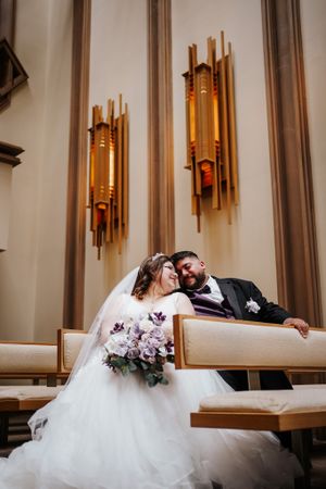 Wedding couple sitting on a church pew with elegant wall sconces and a purple floral bouquet.