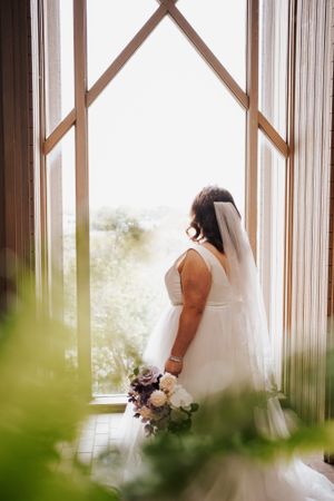 Bride in white dress holding bouquet while standing by geometric window in natural light.