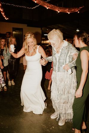 Bride in white gown dances with elderly woman in fringe jacket at wedding reception.