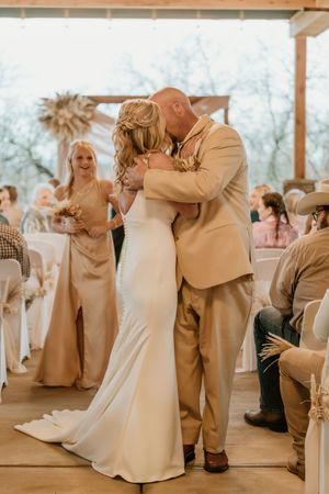 Bride and groom share first kiss at rustic outdoor wedding ceremony with guests and bridesmaid in champagne dress looking on.