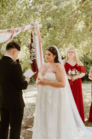 Outdoor wedding ceremony under trees with white arch and red accents.