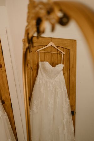 White lace wedding dress hanging on wooden hanger against natural wood door.