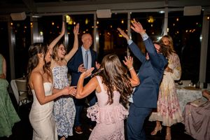 Wedding guests dancing joyfully at a reception, raising hands in celebration under warm string lights.