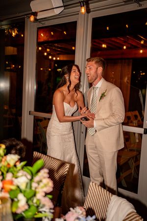 Bride and groom laughing together during wedding reception, bride in white gown, groom in beige suit, string lights behind them.